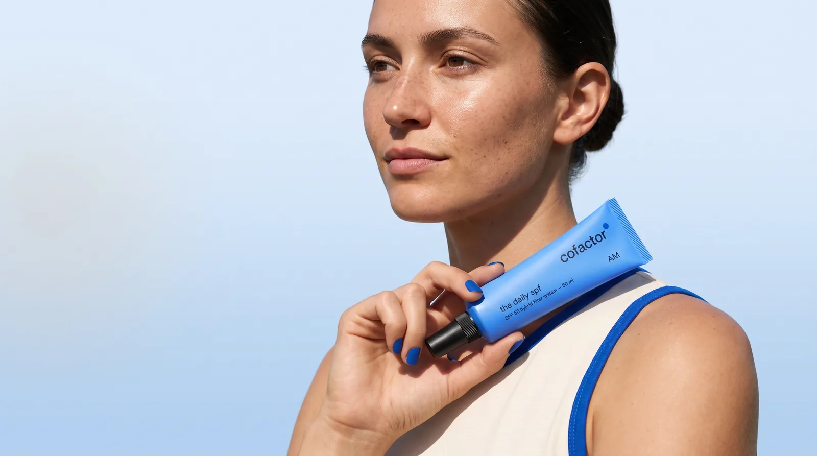 a woman in hard sun against open sky holding the daily spf at her collarbone