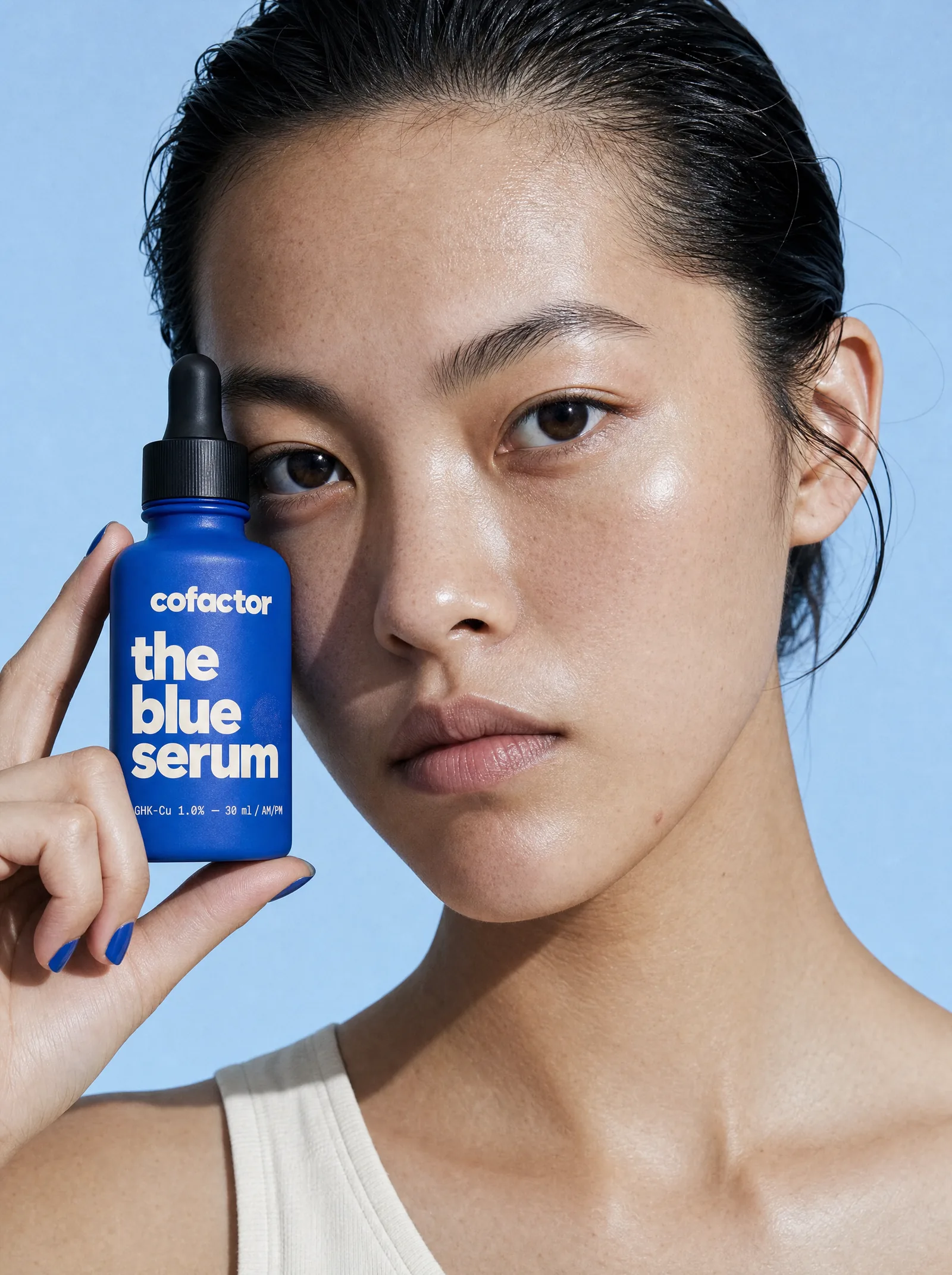 a woman holding the blue serum beside her face, deadpan, in hard sun